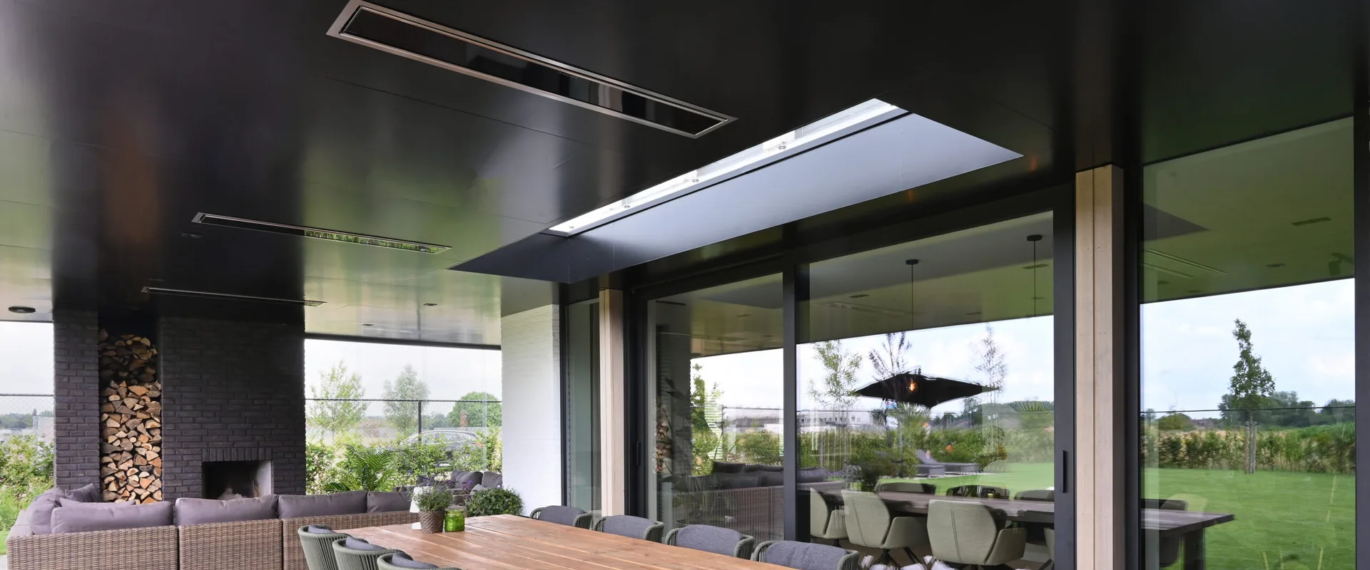 Black Bromic Platinum Electric heaters recessed in the dark ceiling of a covered terrace with a long wooden dining table and garden views