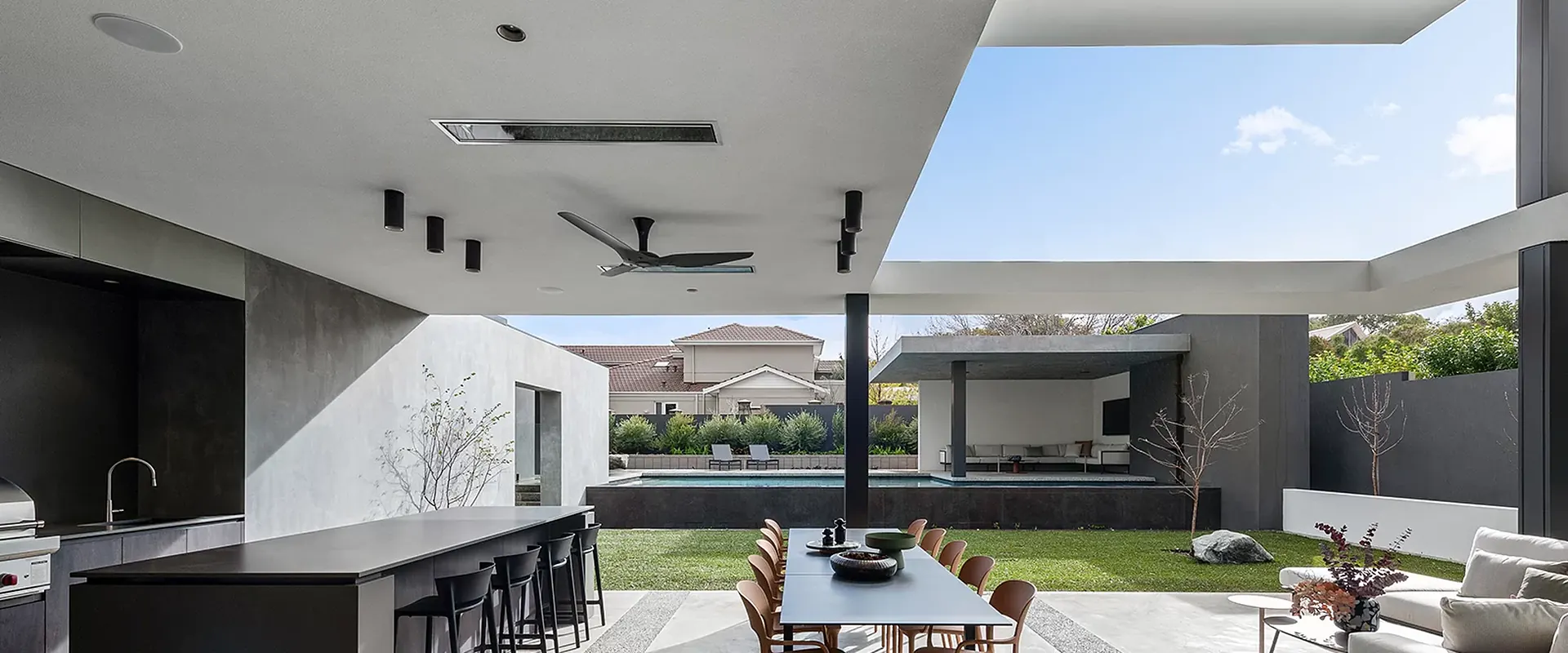 Dark-glass Bromic Platinum Electric heater recessed in the ceiling of a modern concrete terrace with an outdoor kitchen island, a dining table and a pool beyond