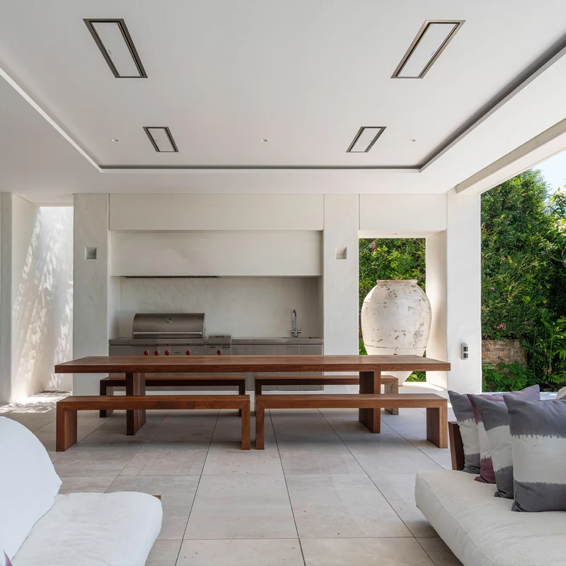 Four Bromic Platinum Electric heaters recessed in a white ceiling above a timber dining table and outdoor kitchen, with a large antique urn and a garden behind