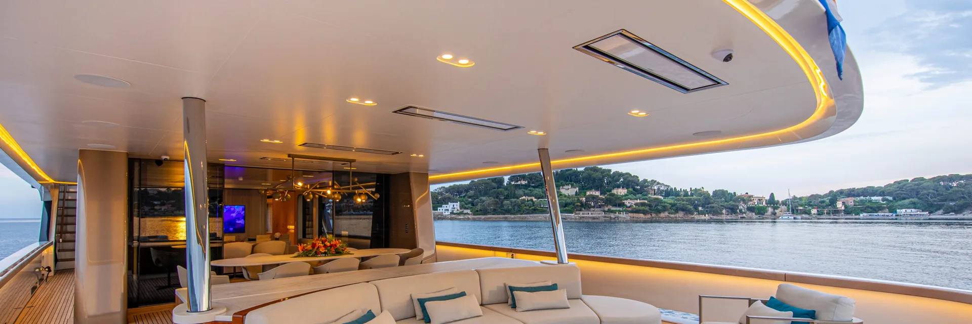 White Bromic Platinum Electric marine heaters recessed in the ceiling above a curved white sofa on a yacht's aft deck, with a wooded coastline behind