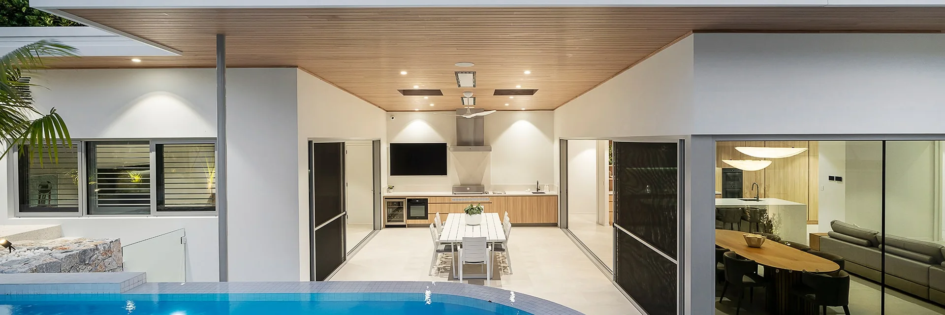 Bromic Platinum Electric heaters recessed in the timber ceiling of an alfresco dining area in a white modern house, with a blue pool in front