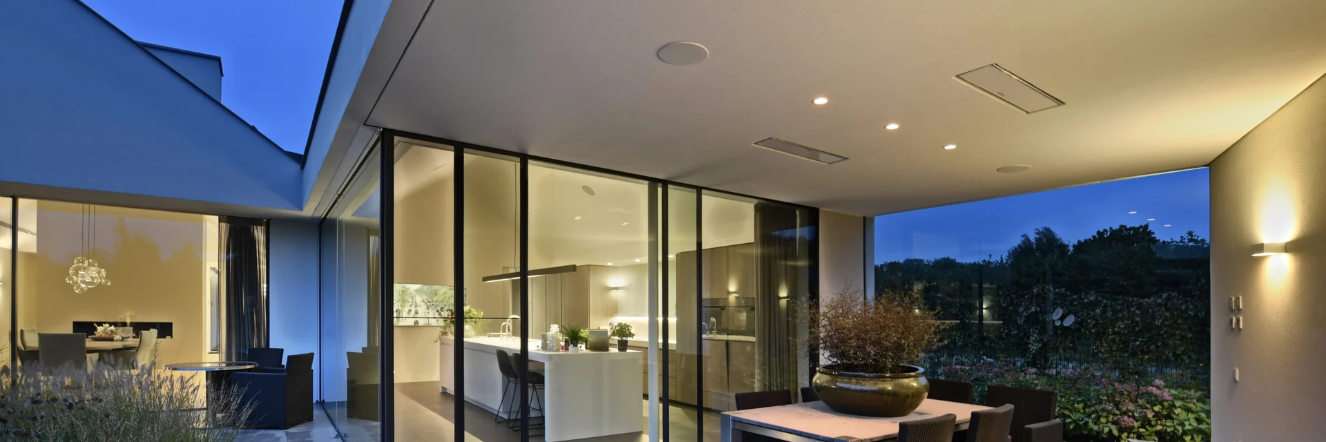 White Bromic Platinum Electric heaters recessed in the ceiling of a covered terrace with a dining table, beside a glass-walled living room at dusk