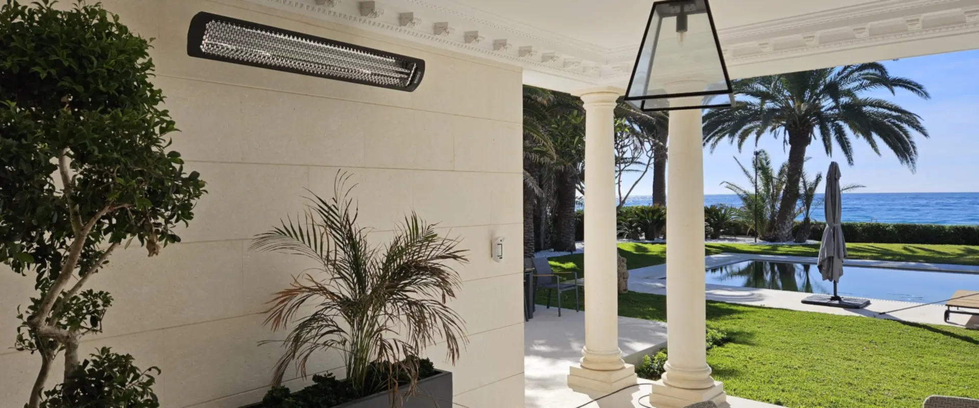 Black 6 kW Bromic Tungsten Electric heater on the stone wall of a covered terrace with columns, palm trees and the sea beyond, Guadalmina