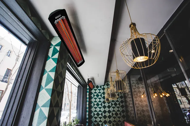 Bromic Tungsten Smart-Heat electric heater glowing on a dark ceiling beside brass cage pendant lamps in a restaurant in Madrid
