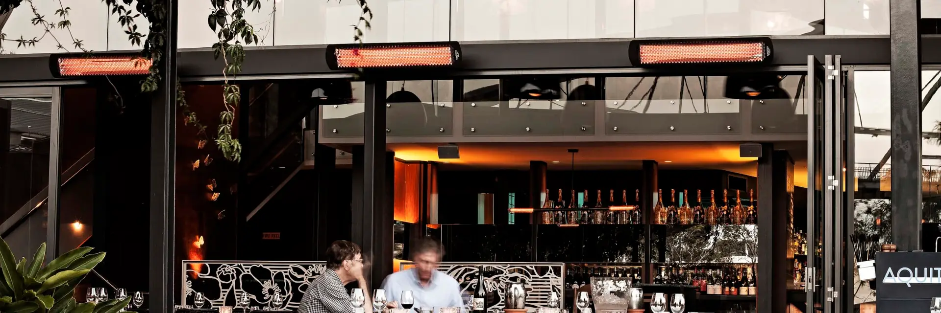 Three Bromic Tungsten Electric heaters glowing above the open front of a restaurant, with diners at a terrace table and a bar behind