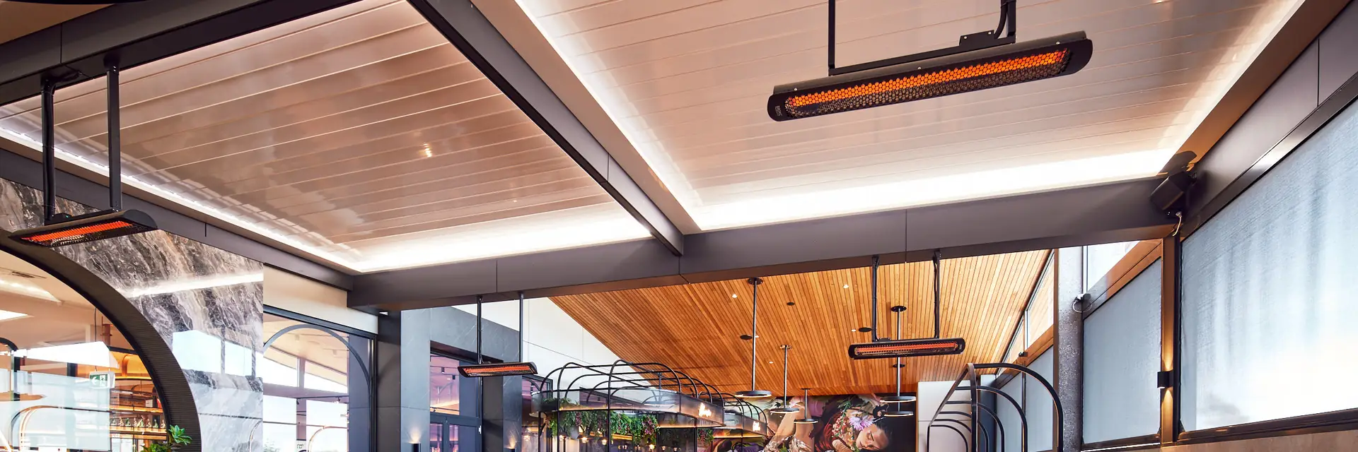 Bromic Tungsten Electric heaters glowing on ceiling poles above the tables of a restaurant terrace with timber ceilings and navy chairs