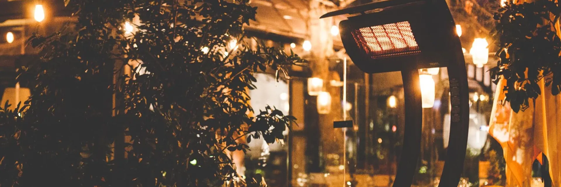 Black Bromic Tungsten Portable gas heater glowing among trees and string lights on a busy restaurant garden terrace at night
