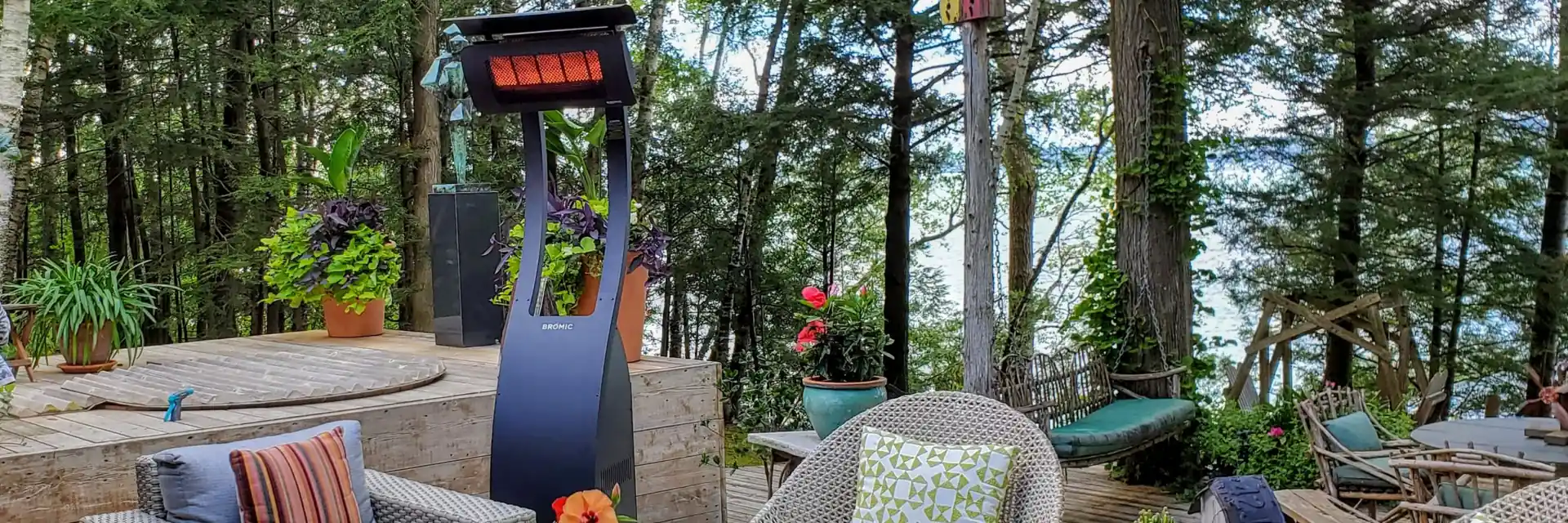 Bromic Tungsten Portable gas heater standing behind wicker armchairs and a wooden coffee table on a patio among tall trees