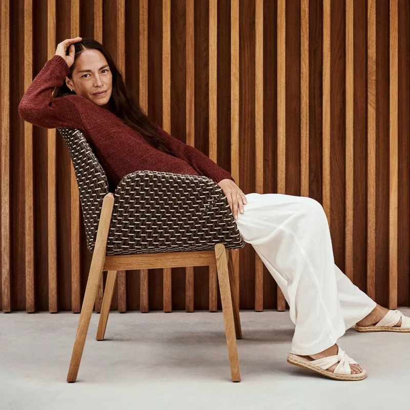 Woman relaxing in a Dedon Caladio armchair with a woven shell and teak legs, in front of a wall of dark timber slats