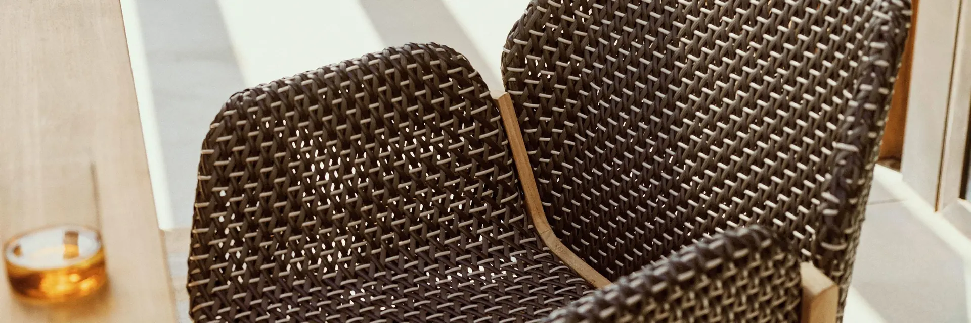 Close view of the dark woven shell and teak legs of a Dedon Caladio armchair beside a teak table