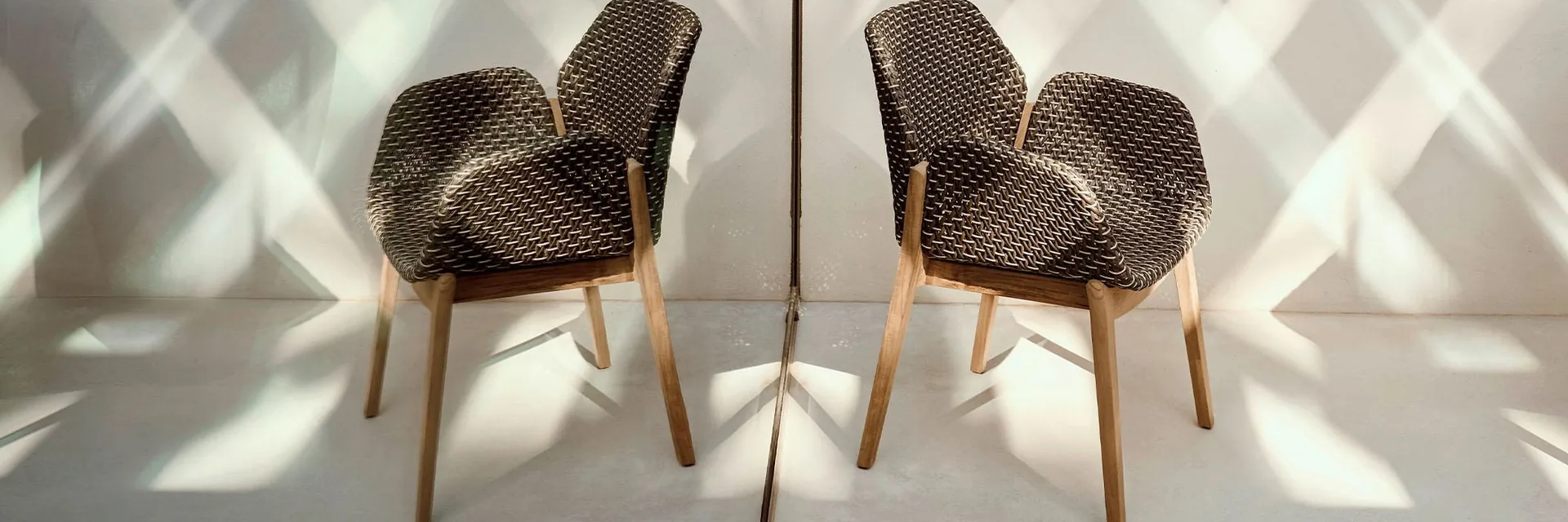 Two Dedon Caladio armchairs with dark woven shells and teak legs facing each other in front of a wall crossed by bands of sunlight