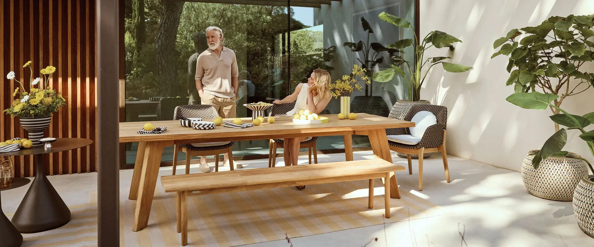 Dedon Caladio woven armchairs at a long teak dining table with a bench on the shaded terrace of a summer house, with two people at the table