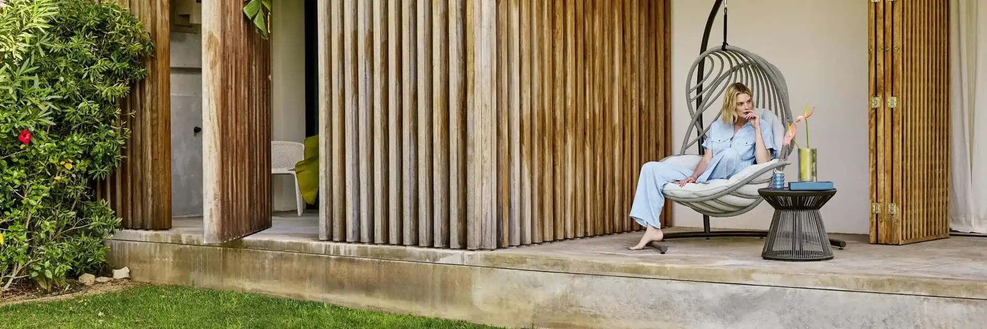 Woman relaxing in a hanging lounge chair beside a Dedon Cirql Nu side table, on the terrace of a timber-clad garden pavilion
