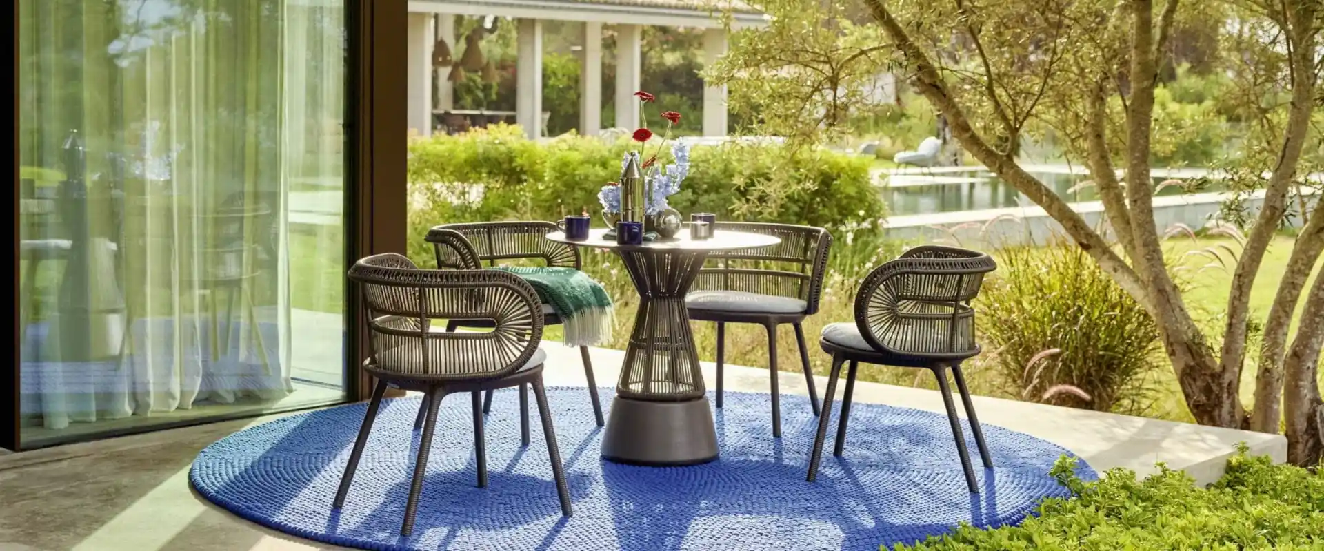 Dedon Cirql Nu woven armchairs around a round bistro table on a blue rug, looking onto a sunny garden