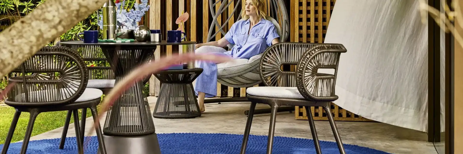 Woman sitting in a Dedon Cirql Nu lounge chair behind a Cirql Nu table and armchairs on a blue rug, in front of a slatted timber screen
