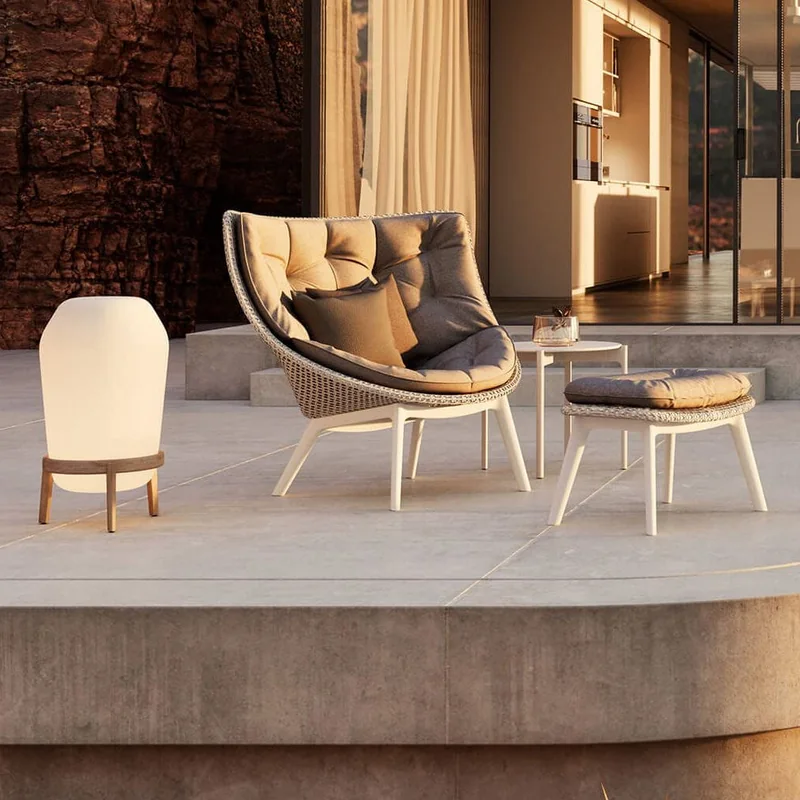 Dedon Mbrace Alu lounge chair with a footstool and a floor lantern on the terrace of a glass-walled house