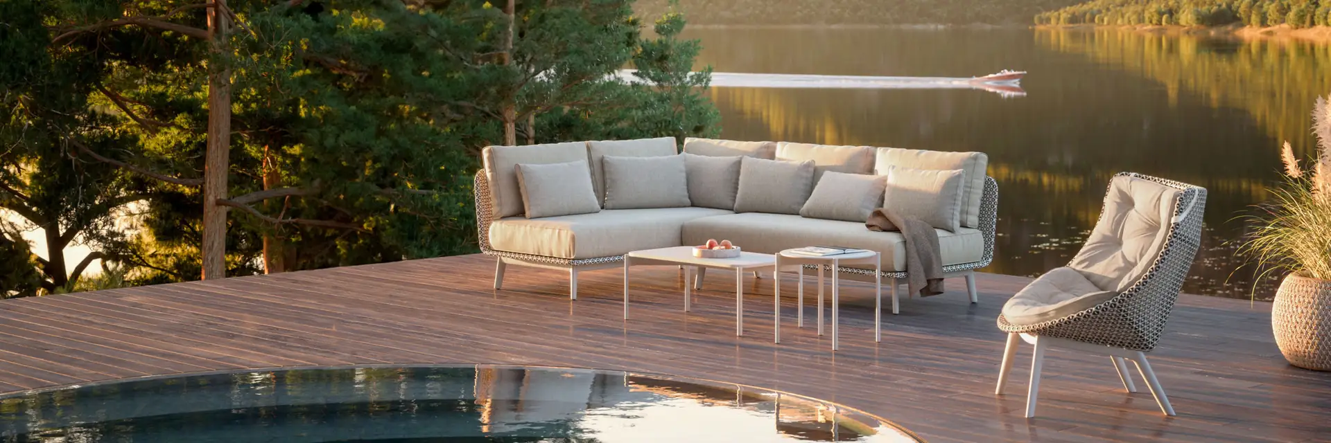 Dedon Mbrace Alu sofa, lounge chair and coffee tables on a timber deck beside a mountain lake at sunset, with pine trees to one side