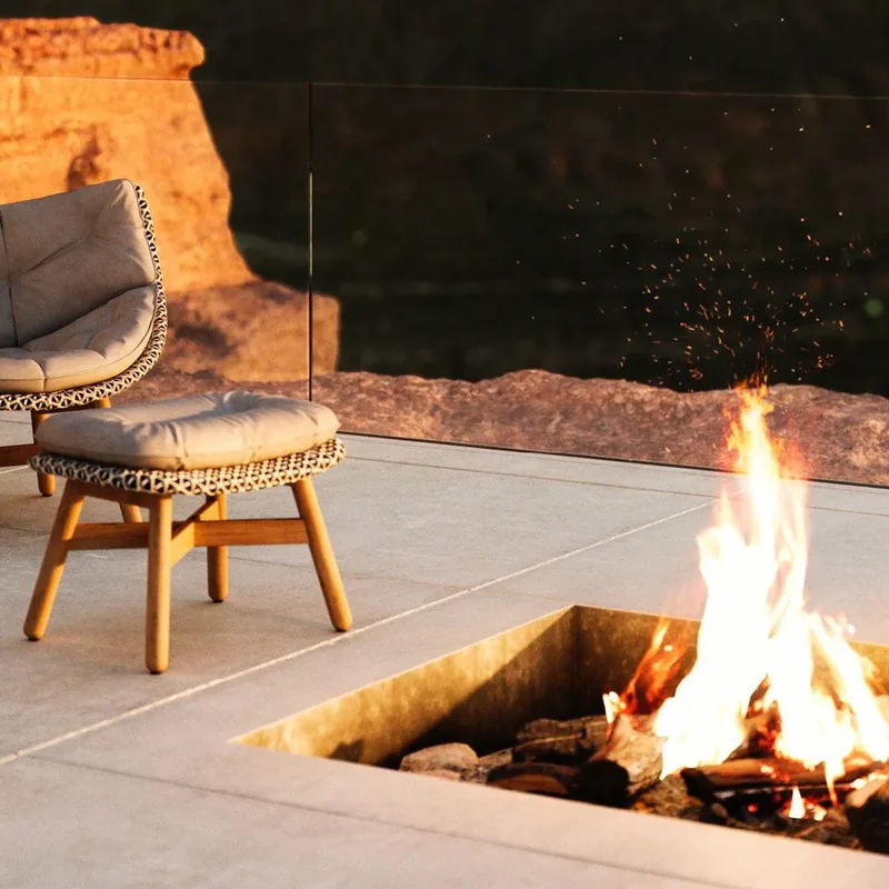 Dedon Mbrace lounge chair with a footstool beside a fire pit on a terrace at dusk