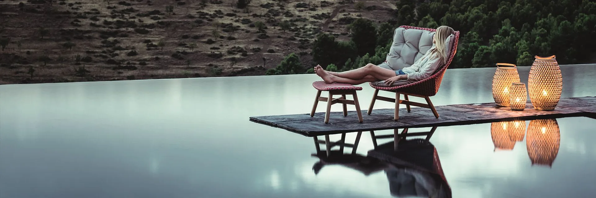 Woman reclining in a Dedon Mbrace lounge chair with a footstool beside an infinity pool at dusk, with lanterns and open heathland behind