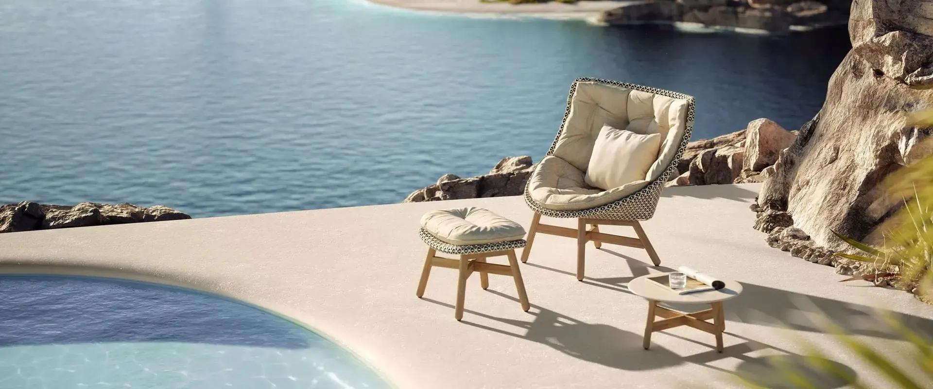Dedon Mbrace lounge chair with a footstool and side table on a pale terrace between a pool and the sea