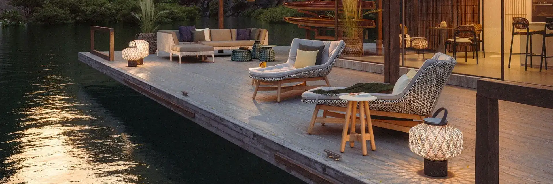 Dedon Mbrace lounge chairs and lanterns on a waterside deck at dusk, in front of a lit timber house