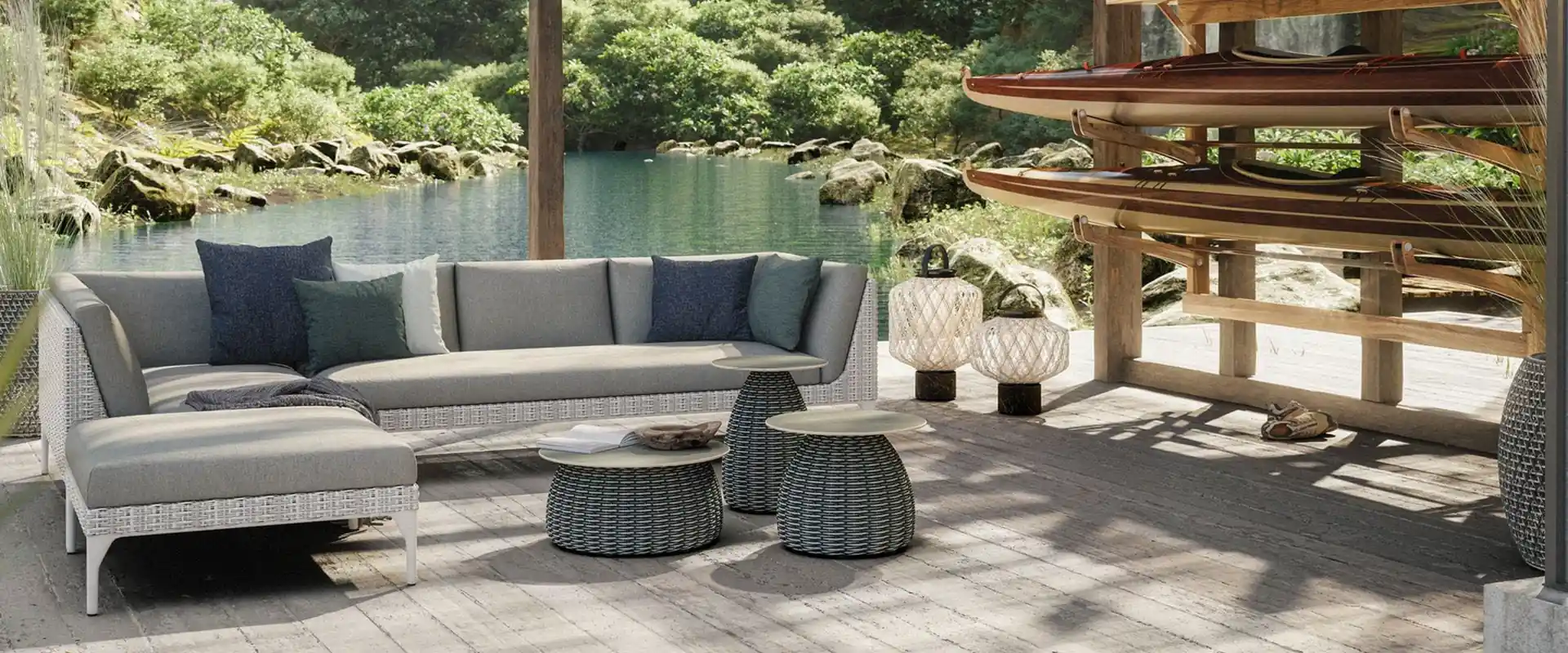 Dedon Mu sofa with blue cushions and woven side tables on a covered deck beside a lake, wooden canoes on a rack