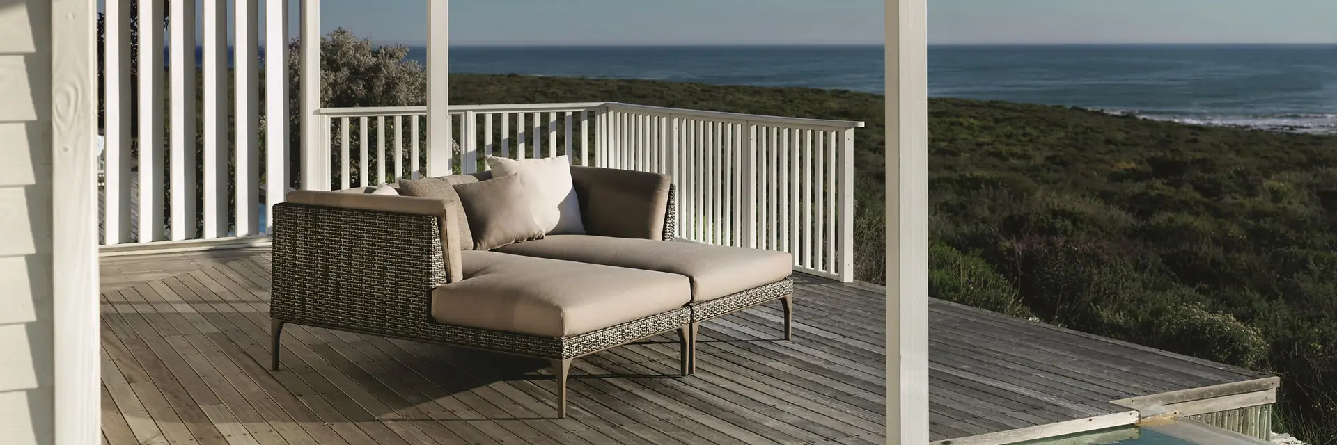 Dedon Mu woven sofa and footstool on the timber deck of a white veranda, with the ocean beyond