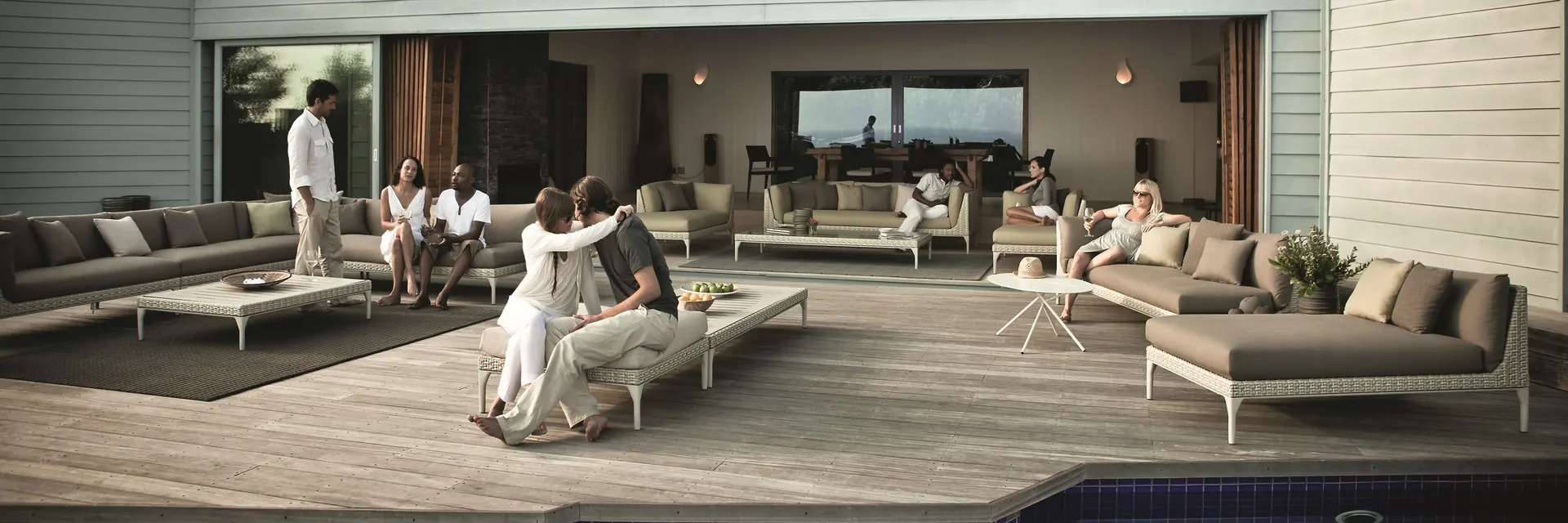Friends relaxing on Dedon Mu sofas and daybeds on the timber terrace of a low modern villa, beside a pool