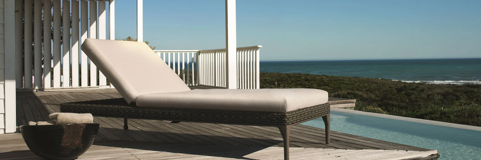 Dedon Mu sun lounger on a timber deck beside a pool, under a white veranda with the ocean on the horizon