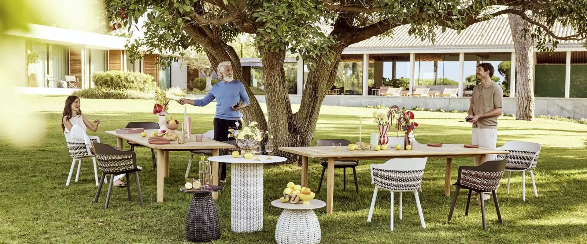 Dedon Mymi chairs with patterned weave around teak tables set for lunch under a large tree in a garden