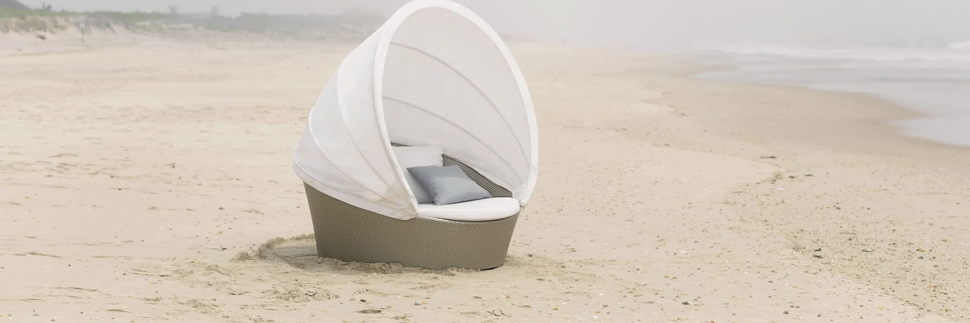 White Dedon Orbit loveseat with its canopy half open on an empty beach in the mist