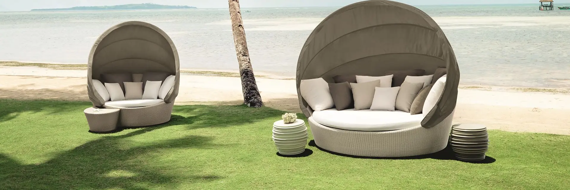 Two Dedon Orbit loveseats with folding canopies on a lawn beside a beach with a leaning palm tree