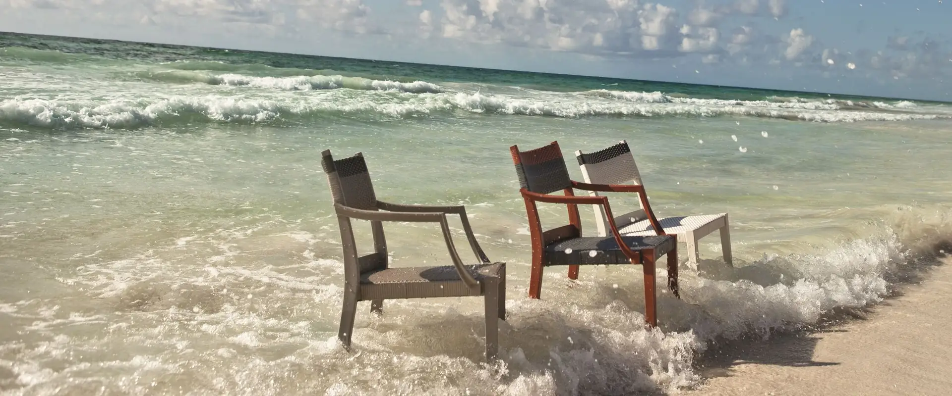 Three Dedon Play armchairs standing in the surf on a tropical beach, under a blue sky with clouds
