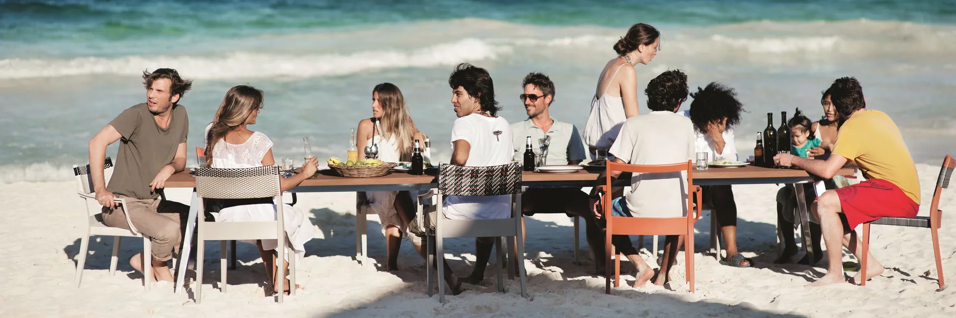 Friends having lunch on Dedon Play chairs at a long table on the sand, with waves behind