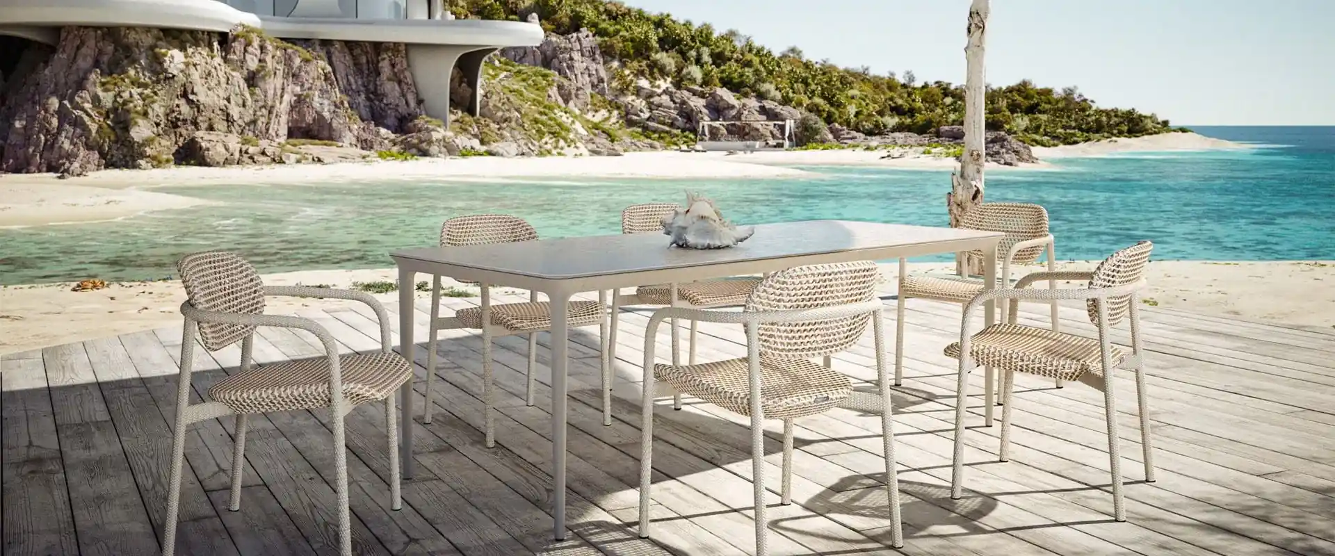 Dedon Roii dining chairs in pale weave around a white table on a wooden deck beside a turquoise bay