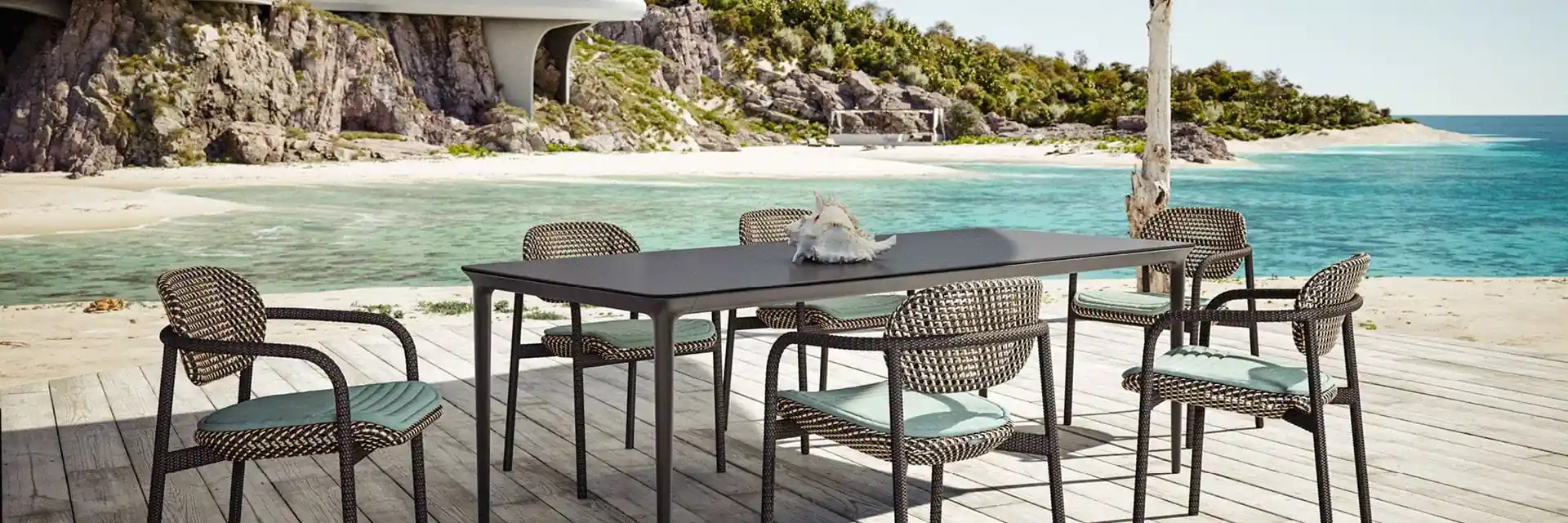 Dedon Roii dining armchairs in dark weave with mint cushions around a long table on a deck above a beach