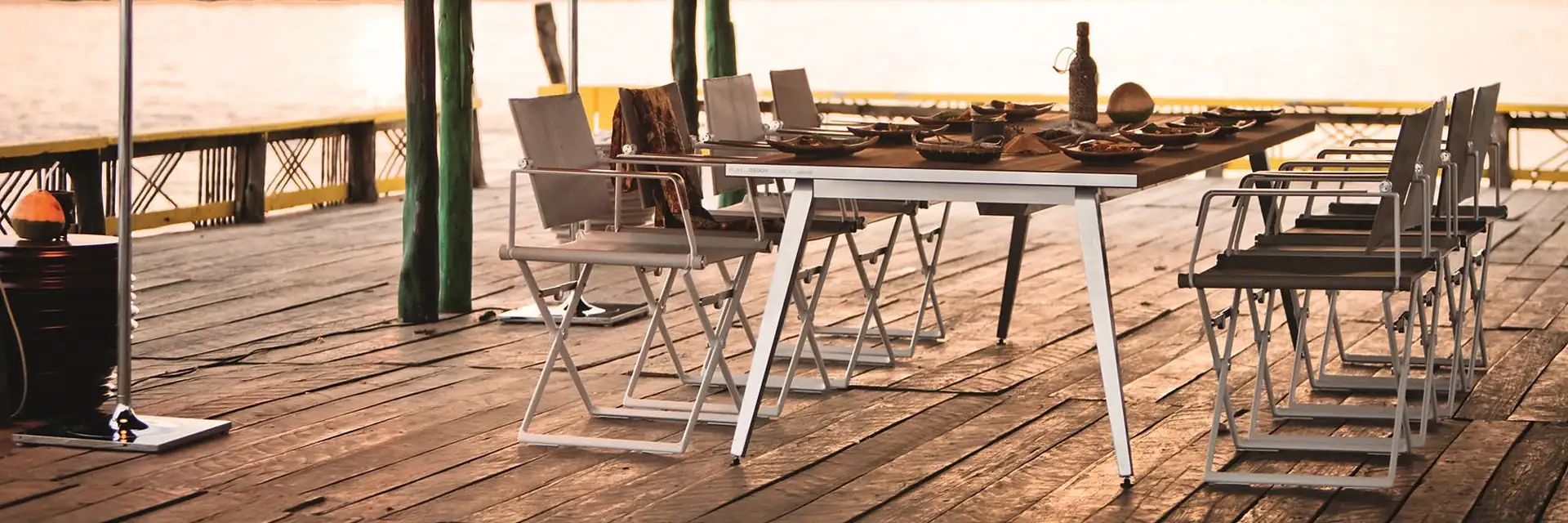 Dedon SEAX armchairs around a long dining table on a timber deck under a thatched roof, beside a lagoon with hills at dusk
