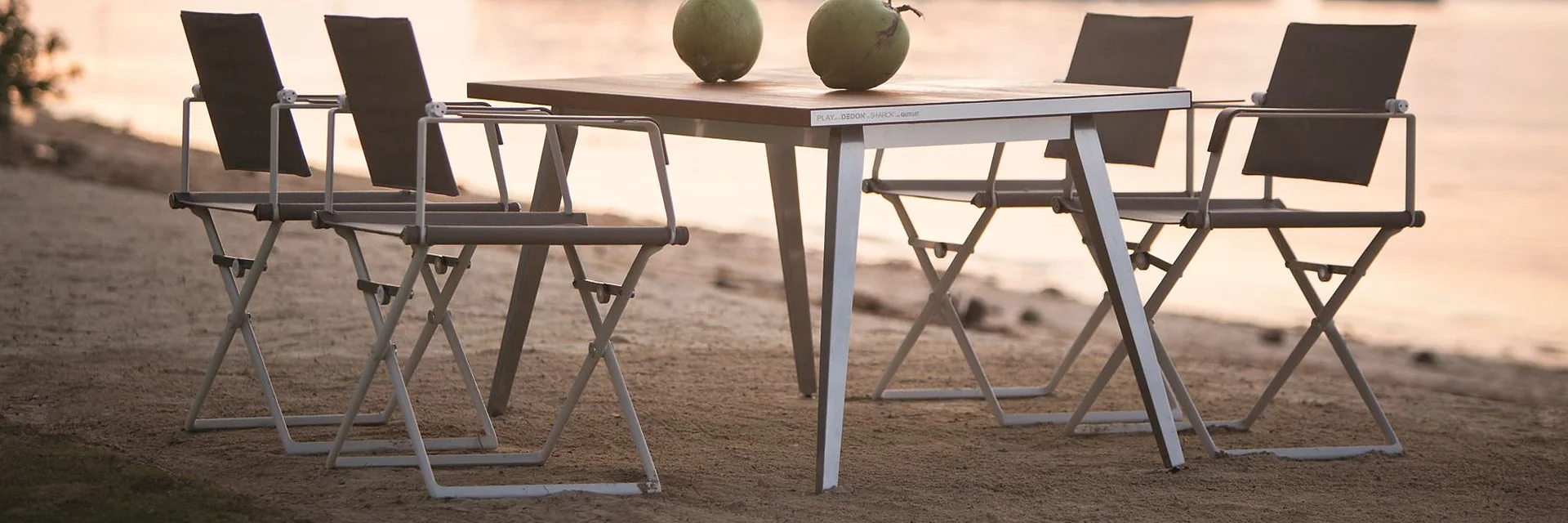 Dedon SEAX folding armchairs around a SEAX dining table on a sandy beach at sunset, with two coconuts on the table and an island offshore