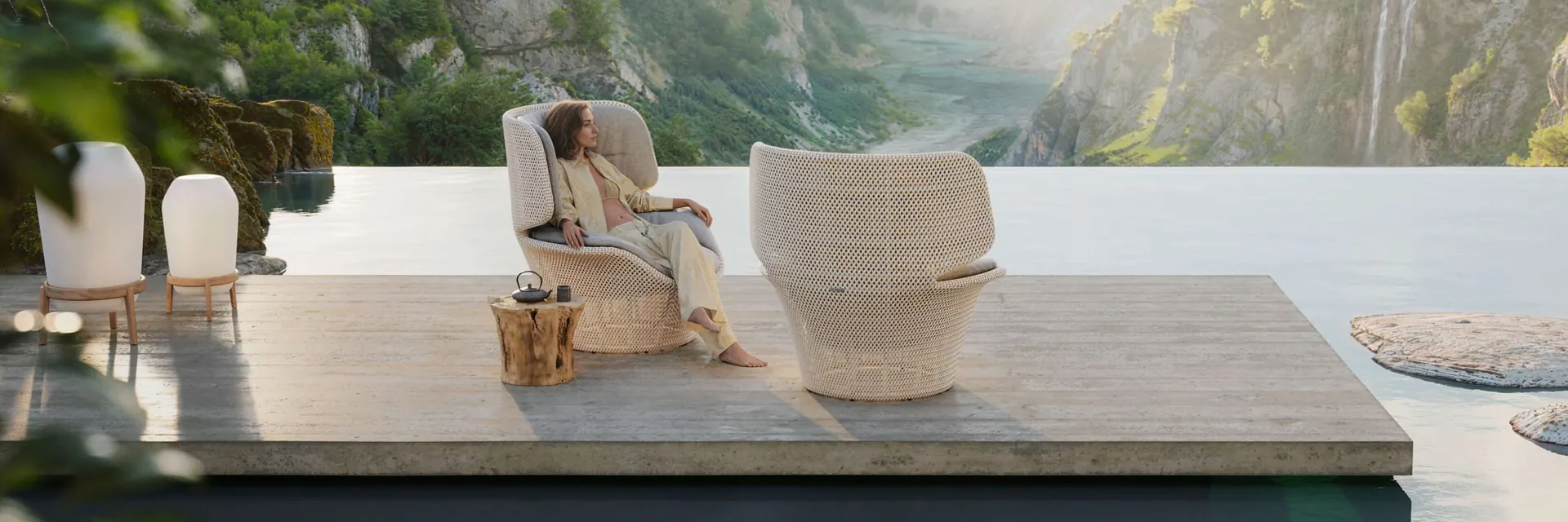 Woman sitting in a Dedon Seashell Grand high-back lounge chair on a stone platform over still water, with steep green mountains behind