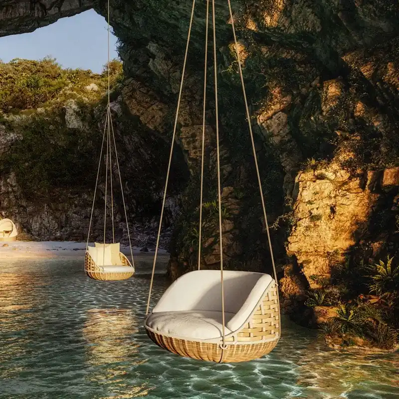Dedon Swingrest hanging lounger suspended over clear shallow water inside a rocky cave