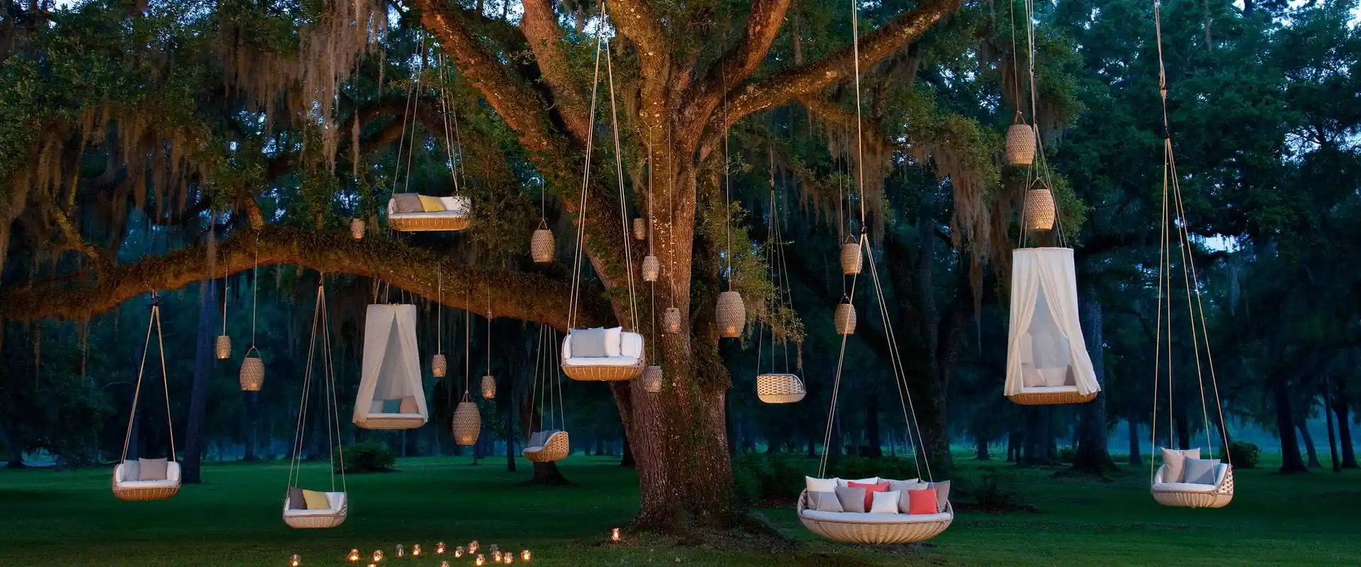 Dedon Swingrest hanging loungers and lanterns suspended from a large tree over a lawn at dusk