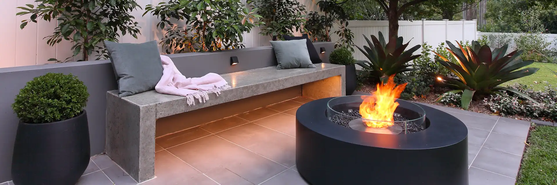 EcoSmart Ark 40 fire table in dark concrete burning beside a long bench in a courtyard garden with trees and potted shrubs