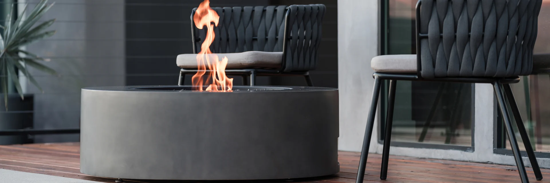 EcoSmart Ark 40 fire table in dark concrete with a tall flame on a timber deck, beside two woven armchairs and a dark modern house
