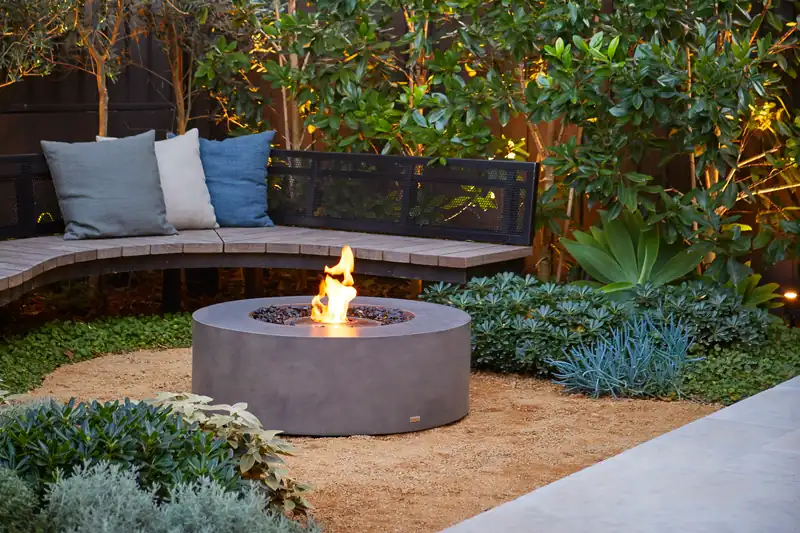 EcoSmart Ark 40 fire table burning in front of a curved bench with cushions in a lush garden at dusk