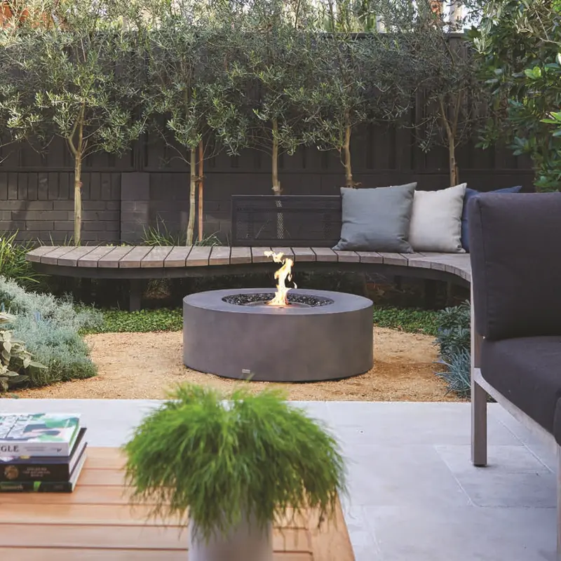 Round dark EcoSmart Ark 40 bioethanol fire table burning on a gravel circle in a landscaped garden, beside an outdoor sofa and a curved timber bench