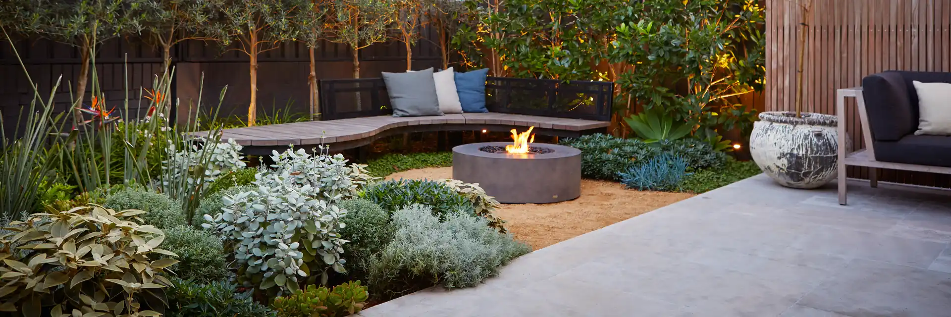 EcoSmart Ark 40 fire table in pale concrete burning in a landscaped garden with a low bench, grasses and trees