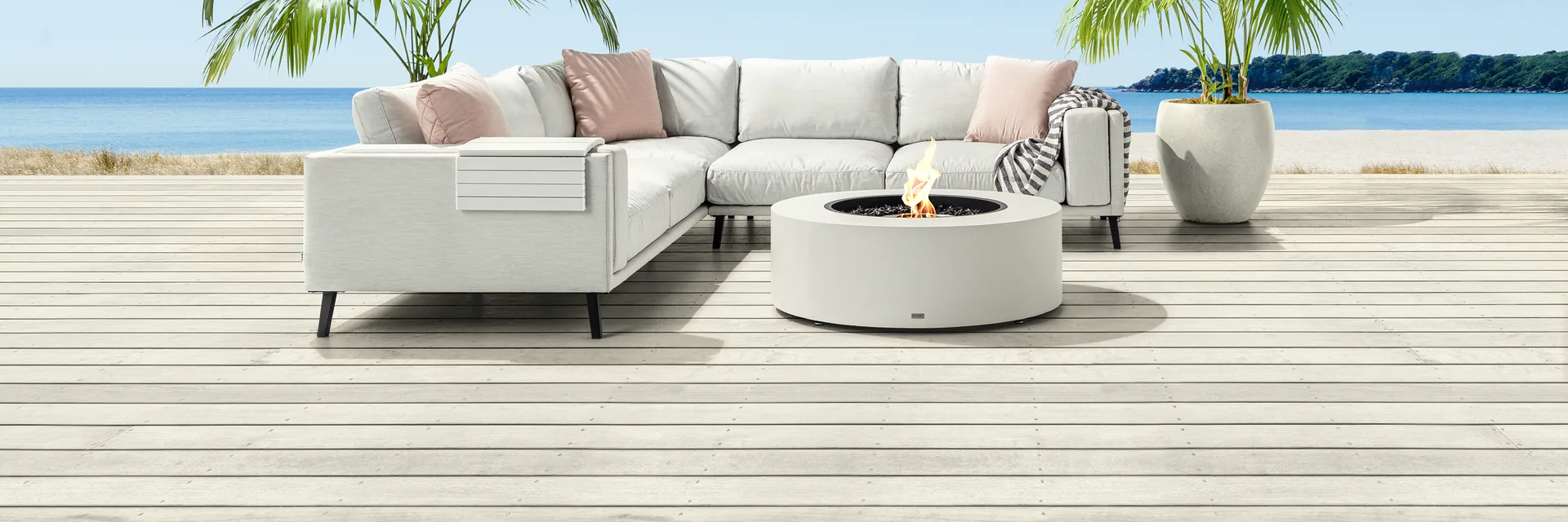 EcoSmart Ark 40 fire table in white beside a white corner sofa on a pale deck, with palm trees and the sea behind