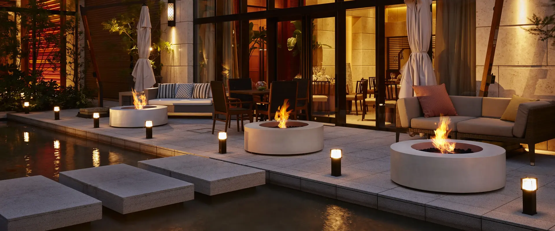 Three round pale EcoSmart Ark 40 bioethanol fire tables burning along a stone terrace beside a pond at night, with lounge seating and a glass-fronted restaurant behind