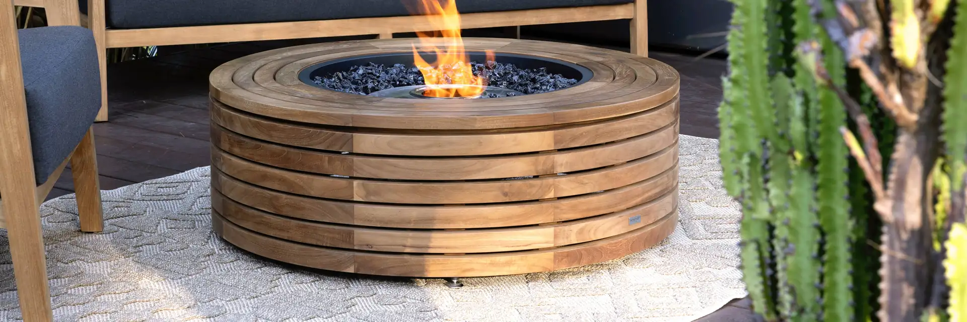 EcoSmart Ark 40 fire table in teak burning in front of an outdoor sofa with tropical-print cushions, beside a tall cactus