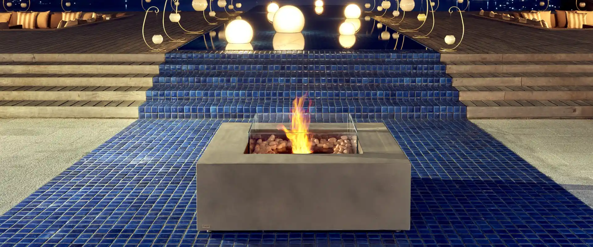 Square concrete EcoSmart Base 40 bioethanol fire table with a glass screen burning on a blue mosaic terrace at dusk, below steps lined with glowing lamps and a view of distant hills